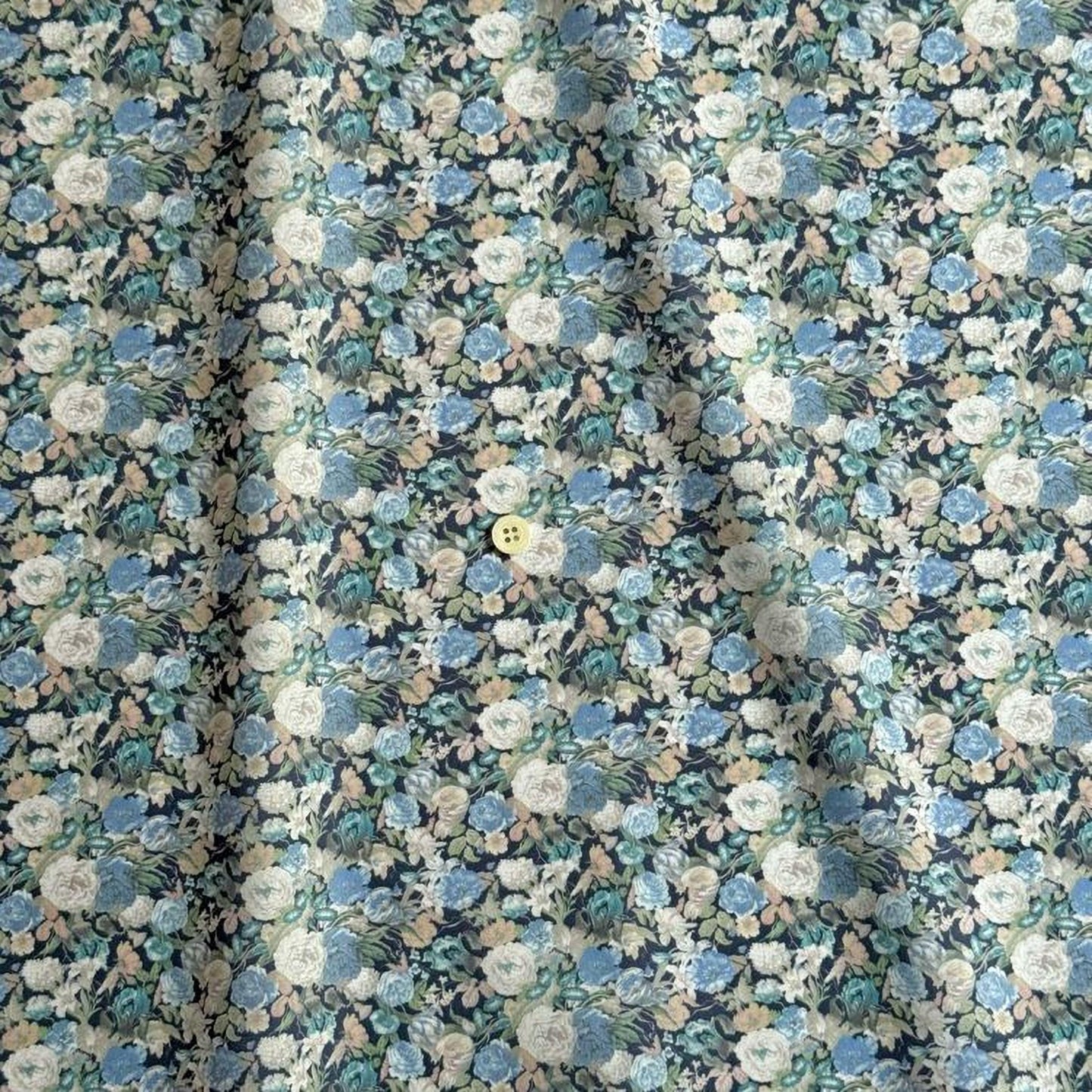 a close-up view of a fabric with a floral pattern, featuring a variety of colors including blue, green, and white. The fabric appears to be made of a soft, textured material, and there is a small yellow button visible in the center of the pattern.