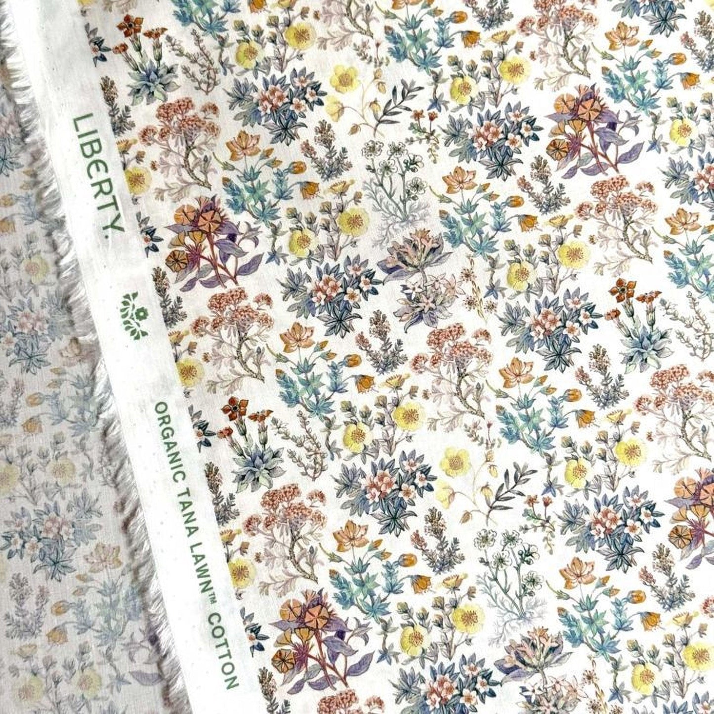 a close-up view of a fabric with a floral pattern. The fabric appears to be a white material with a variety of colorful flowers and leaves printed on it. The text "LIBERTY" and "ORGANIC TAN A LEW'N COTON" can be seen, indicating the type of fabric and its origin.