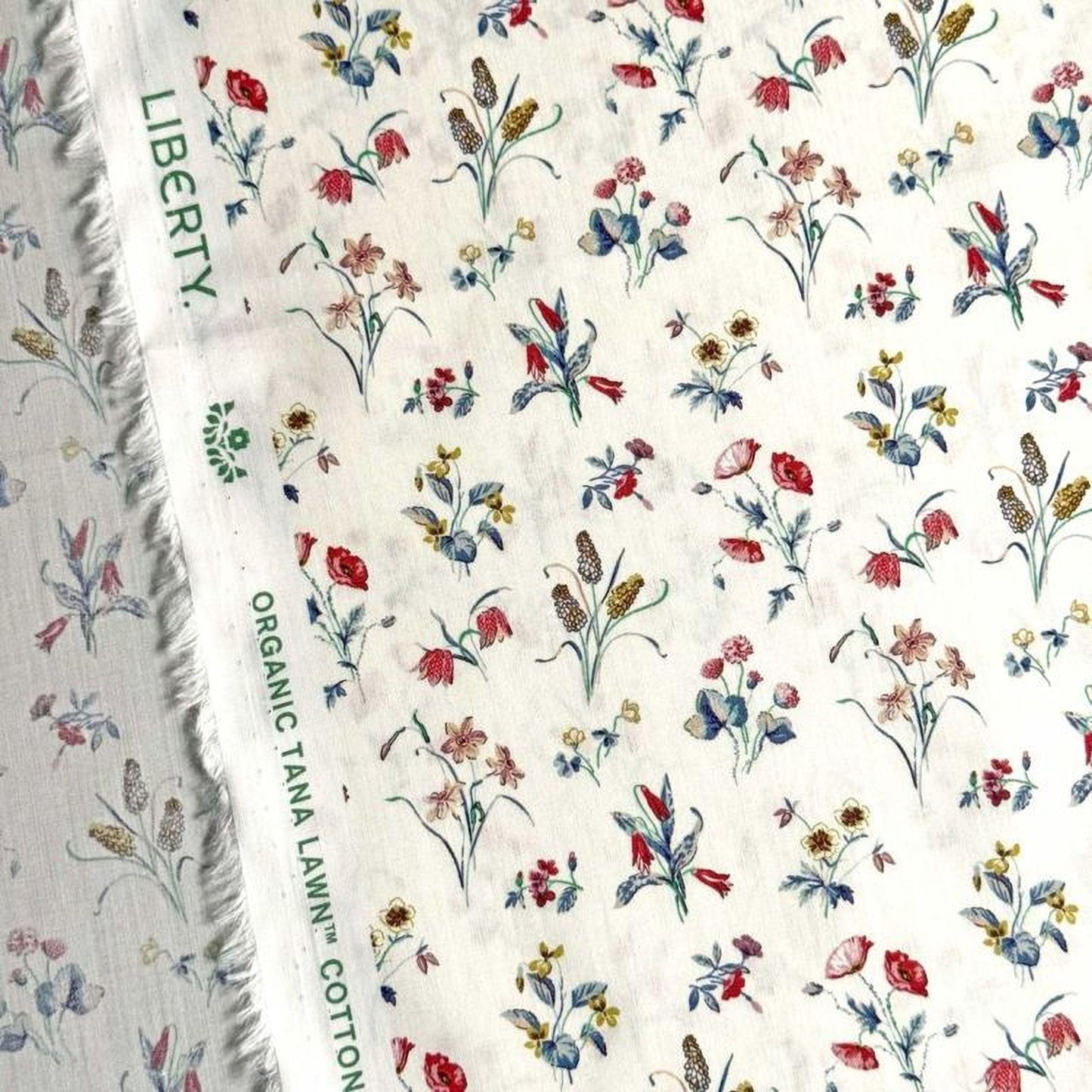 a close-up view of a fabric with a floral pattern, featuring various types of flowers and leaves in different colors. The fabric appears to be a white material with a green label that reads "Liberty Organic Tana Lawn Cotton".