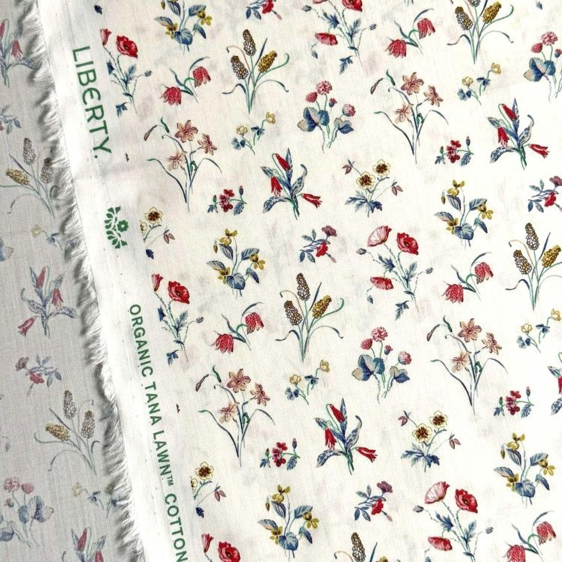 a close-up view of a fabric with a floral pattern, featuring various types of flowers and leaves in different colors. The fabric appears to be a white material with a green label that reads "Liberty Organic Tana Lawn Cotton".