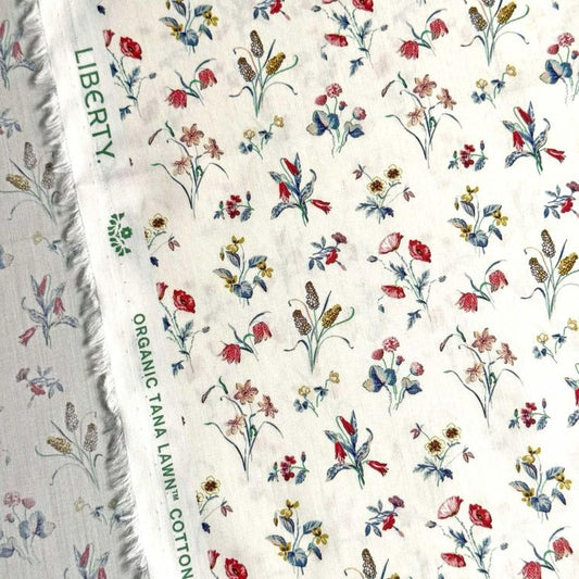 a close-up view of a fabric with a floral pattern, featuring various types of flowers and leaves in different colors. The fabric appears to be a white material with a green label that reads "Liberty Organic Tana Lawn Cotton".