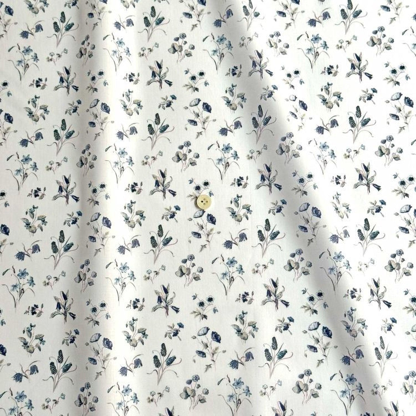 a close-up view of a fabric with a repeating floral pattern in shades of blue and white. The pattern consists of various flowers and leaves, creating a visually appealing and intricate design. There is a single button visible in the center of the fabric, adding a contrasting element to the overall pattern.