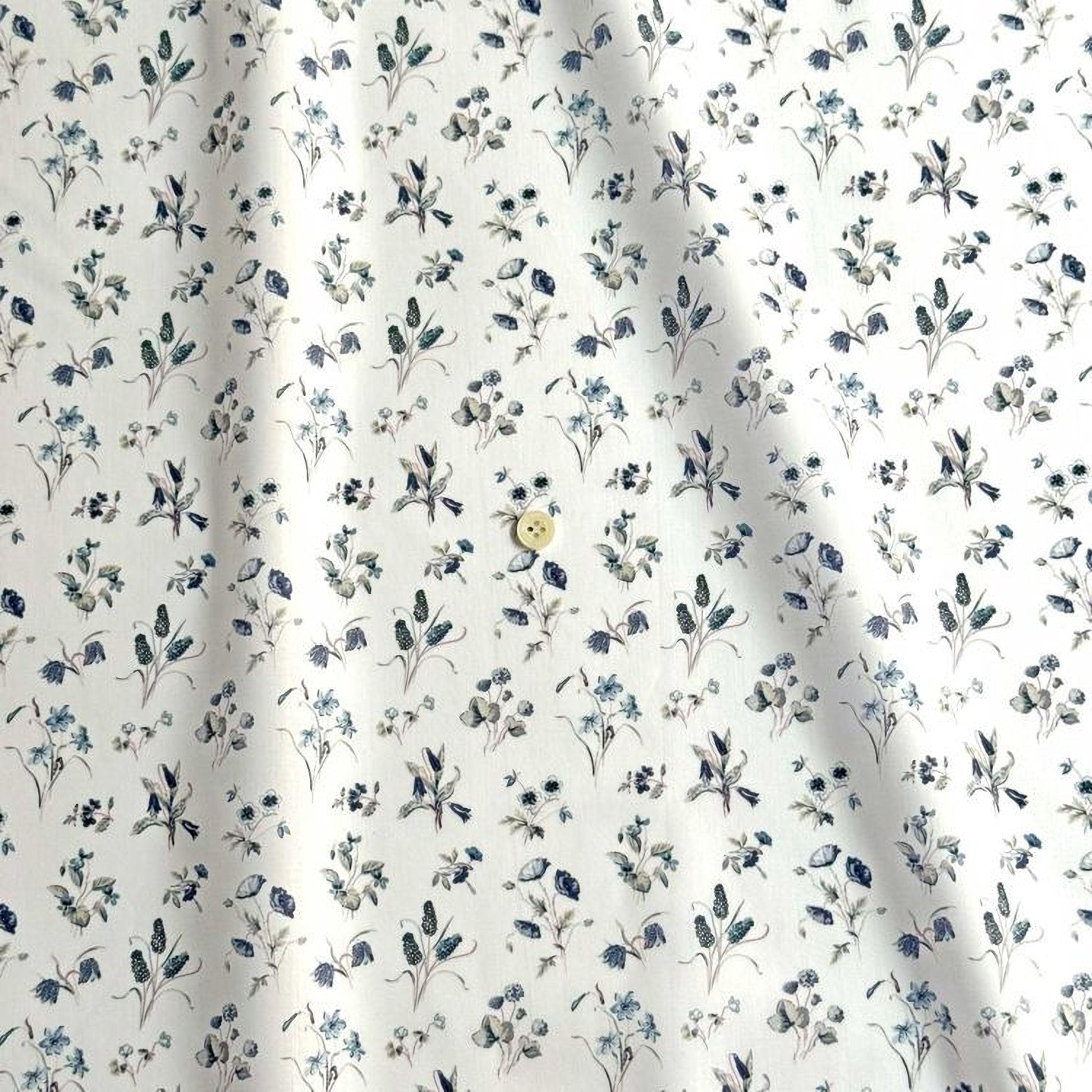 a close-up view of a fabric with a repeating floral pattern in shades of blue and white. The pattern consists of various flowers and leaves, creating a visually appealing and intricate design. There is a single button visible in the center of the fabric, adding a contrasting element to the overall pattern.