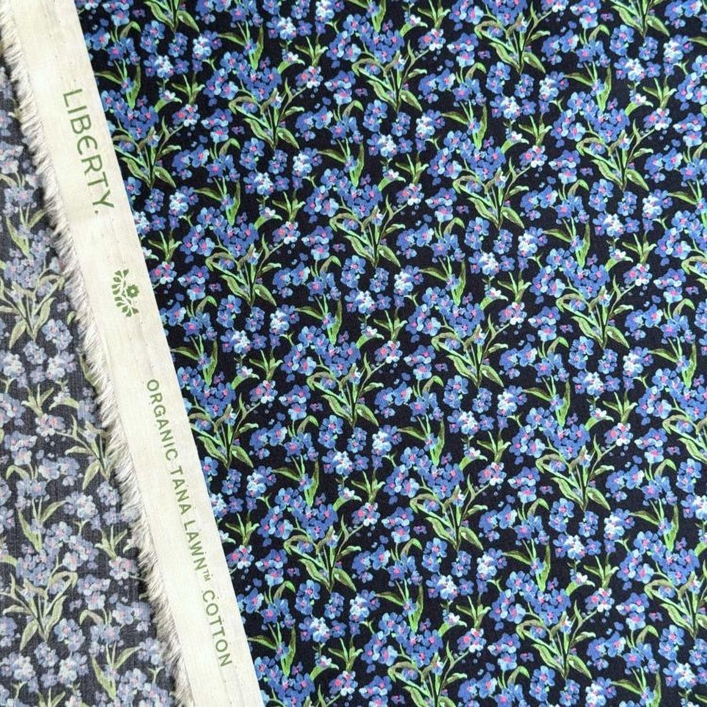 a close-up view of a fabric with a floral pattern in shades of blue and green. The fabric appears to be a blend of black and white, with the blue and green colors standing out against the black background. The fabric is described as "Liberty" and "Organic Tana Lawn Cotton", indicating that it is a high-quality, organic cotton fabric with a floral print design.