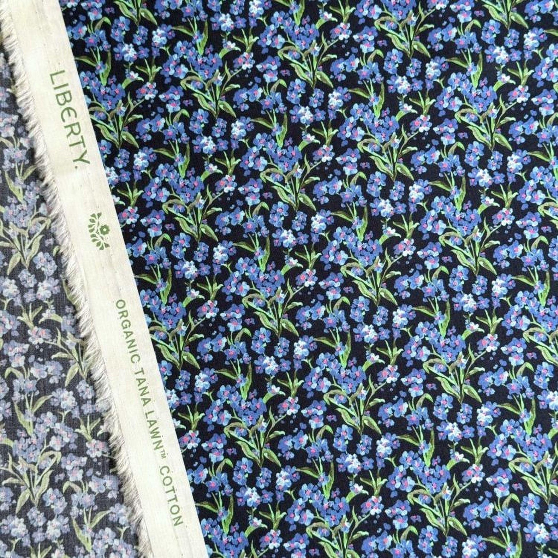 a close-up view of a fabric with a floral pattern in shades of blue and green. The fabric appears to be a blend of black and white, with the blue and green colors standing out against the black background. The fabric is described as "Liberty" and "Organic Tana Lawn Cotton", indicating that it is a high-quality, organic cotton fabric with a floral print design.