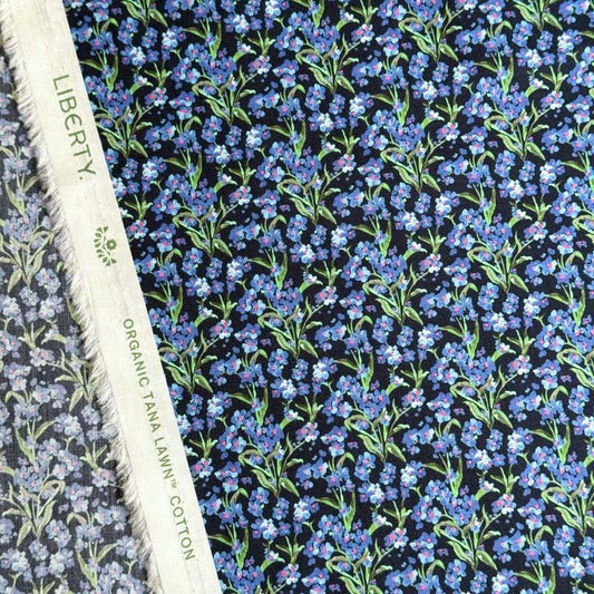a close-up view of a fabric with a floral pattern in shades of blue and green. The fabric appears to be a blend of black and white, with the blue and green colors standing out against the black background. The fabric is described as "Liberty" and "Organic Tana Lawn Cotton", indicating that it is a high-quality, organic cotton fabric with a floral print design.