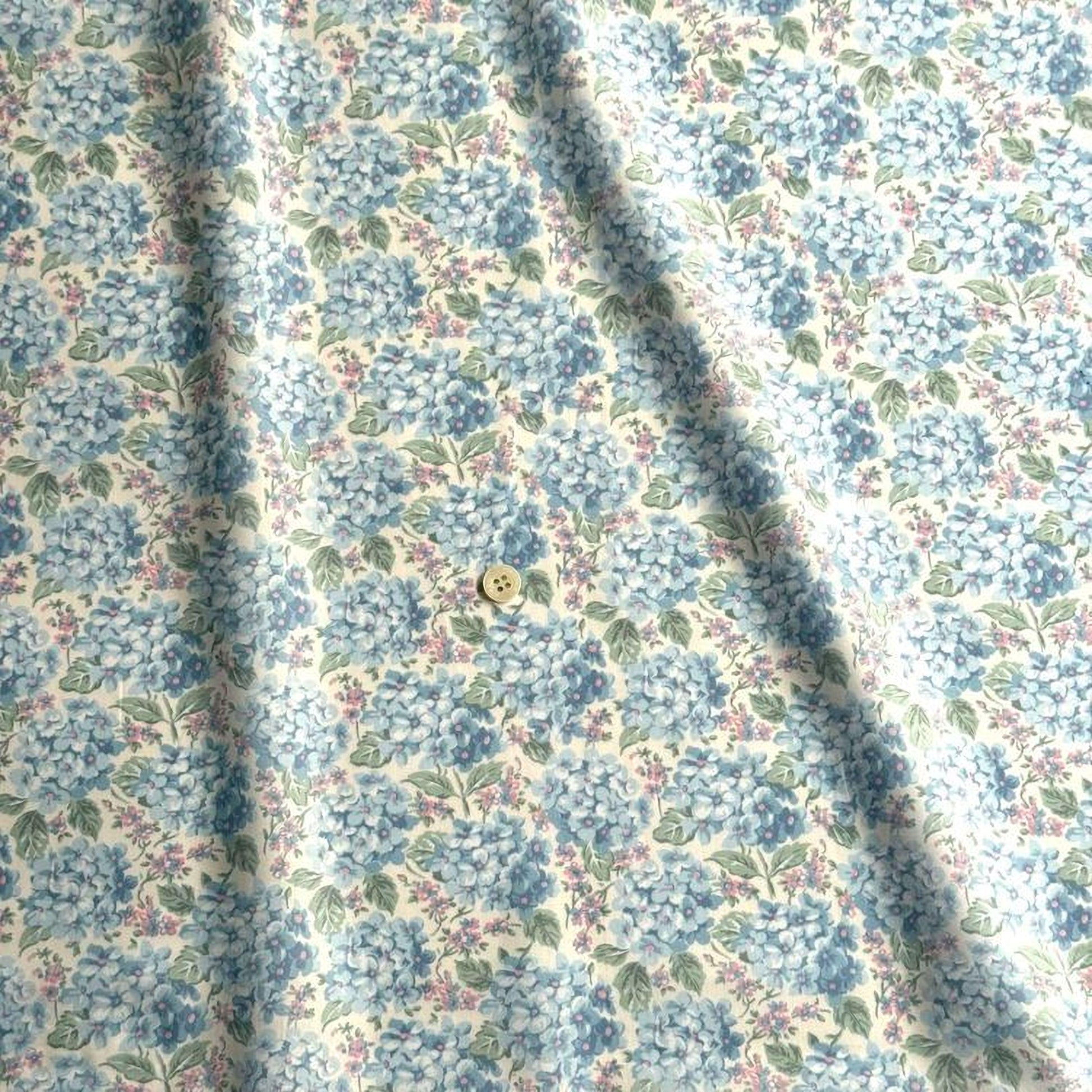 a close-up view of a fabric with a floral pattern, featuring blue hydrangea flowers and green leaves. There is a button visible in the center of the fabric.
