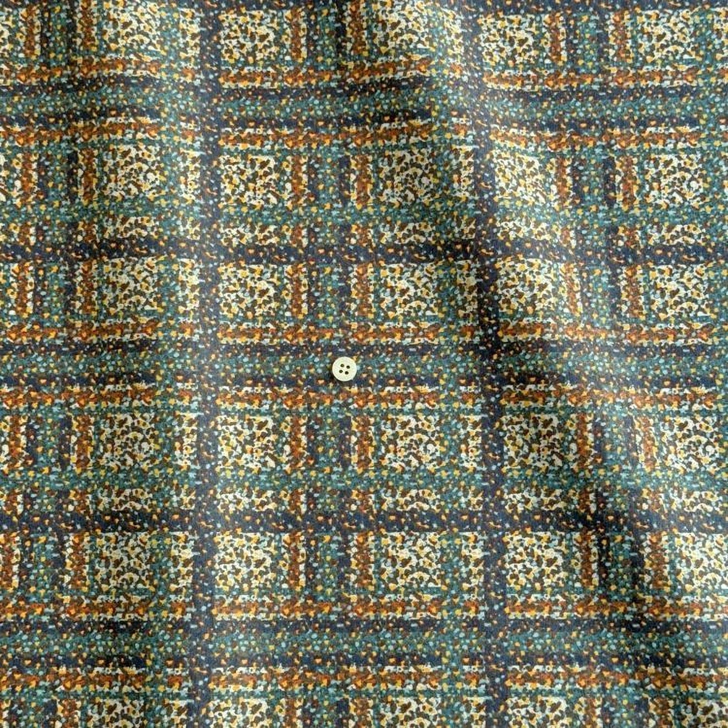 a close-up view of a fabric with a colorful, abstract pattern of squares and rectangles in various shades of green, orange, and brown. There is a small white button visible in the center of the pattern.