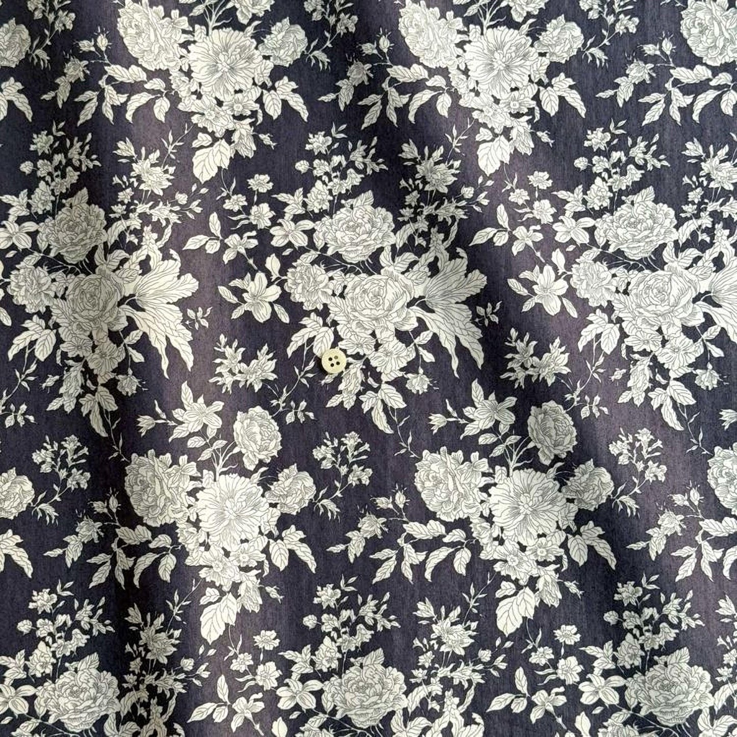 a close-up view of a fabric with a floral pattern in shades of gray and white. The pattern consists of numerous white flowers and leaves, creating a visually striking and intricate design. The fabric appears to be dark gray, providing a contrast to the lighter floral elements. The image does not contain any text or additional objects, and the focus is solely on the floral pattern.