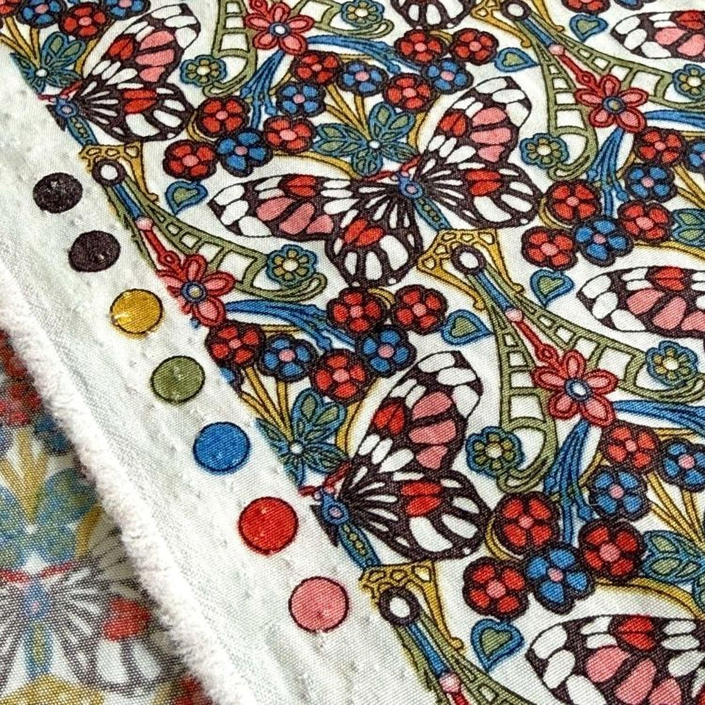 a close-up view of a vibrant, colorful fabric with a repeating pattern of butterflies and flowers. The fabric appears to be a textile, possibly a piece of clothing or a decorative item. The pattern is intricate and detailed, with the butterflies and flowers rendered in a variety of colors, including red, blue, green, and yellow. The fabric is adorned with several small circles, which are likely part of the design or embellishments.