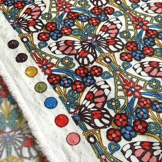a close-up view of a vibrant, colorful fabric with a repeating pattern of butterflies and flowers. The fabric appears to be a textile, possibly a piece of clothing or a decorative item. The pattern is intricate and detailed, with the butterflies and flowers rendered in a variety of colors, including red, blue, green, and yellow. The fabric is adorned with several small circles, which are likely part of the design or embellishments.