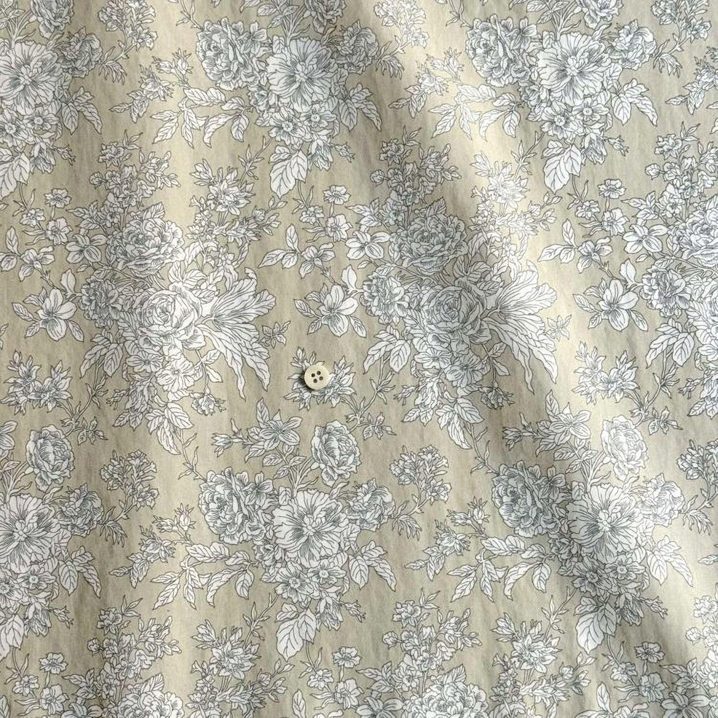 a close-up view of a fabric with a floral pattern. The fabric appears to be a light-colored material with a repeating floral design in shades of gray and white. There is a small button visible in the center of the fabric.