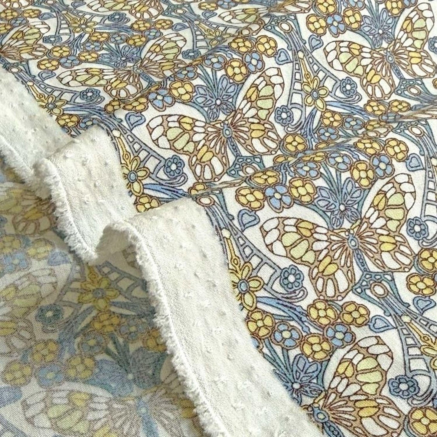 a close-up view of a fabric with a floral pattern, including yellow and blue flowers, butterflies, and other decorative elements. The fabric appears to be a piece of cloth with a fringed edge, and the background is blurred, drawing attention to the intricate floral design.