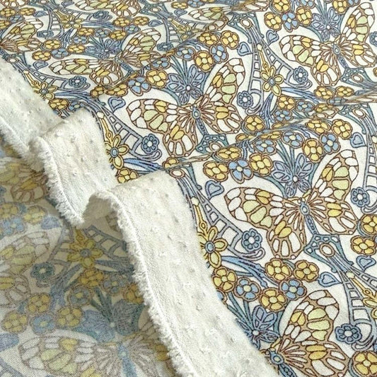 a close-up view of a fabric with a floral pattern, including yellow and blue flowers, butterflies, and other decorative elements. The fabric appears to be a piece of cloth with a fringed edge, and the background is blurred, drawing attention to the intricate floral design.