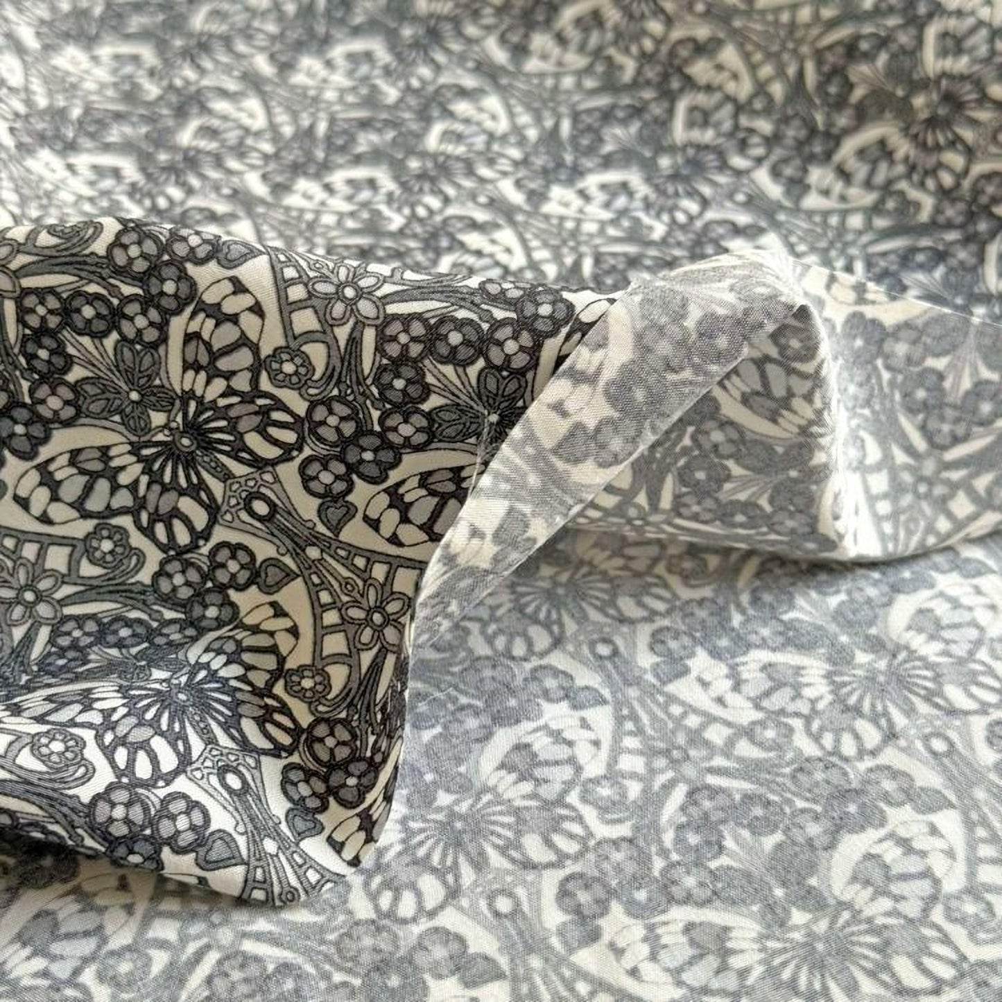 a close-up view of a fabric with a repeating floral pattern in shades of gray and white. The pattern appears to be a printed design, with intricate details and a textured appearance.