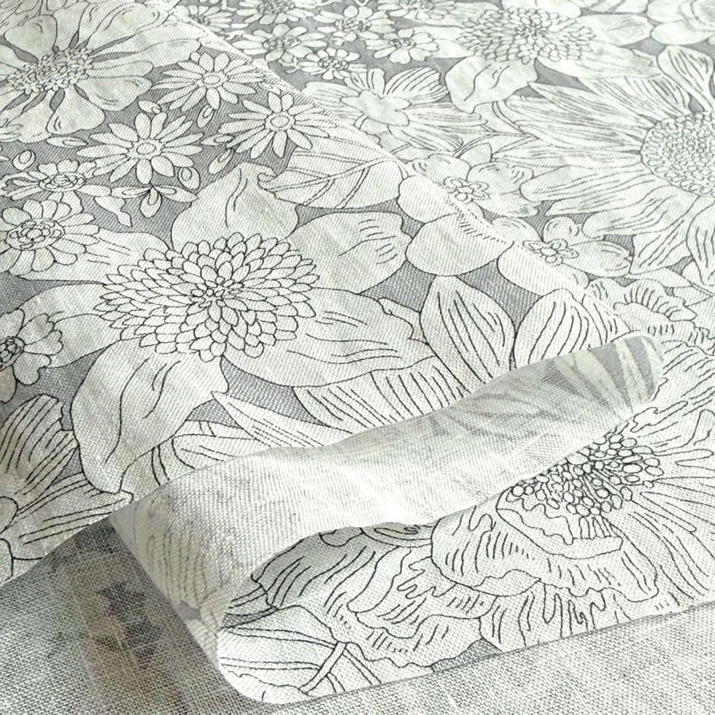 a close-up view of a fabric with a floral pattern, including various types of flowers and leaves. The fabric appears to be a piece of linens or a textile, with a gray and white color scheme.