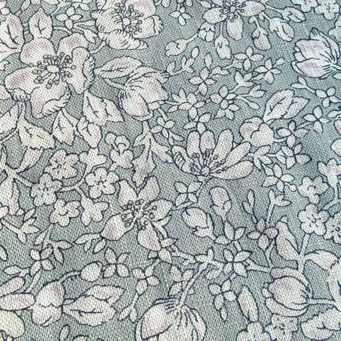 a close-up view of a fabric with a floral pattern. The fabric appears to be a light blue color, and the pattern consists of various flowers and leaves in shades of white and gray. The image does not contain any text or other discernible objects or actions.