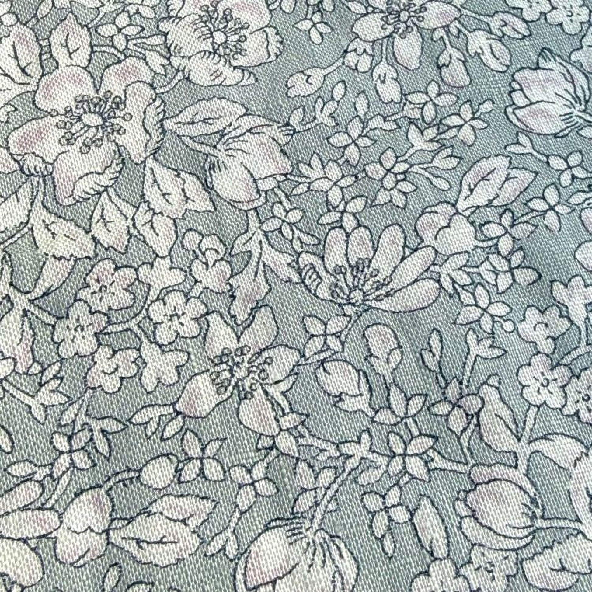 a close-up view of a fabric with a floral pattern. The fabric appears to be a light blue color, and the pattern consists of various flowers and leaves in shades of white and gray. The image does not contain any text or other discernible objects or actions.