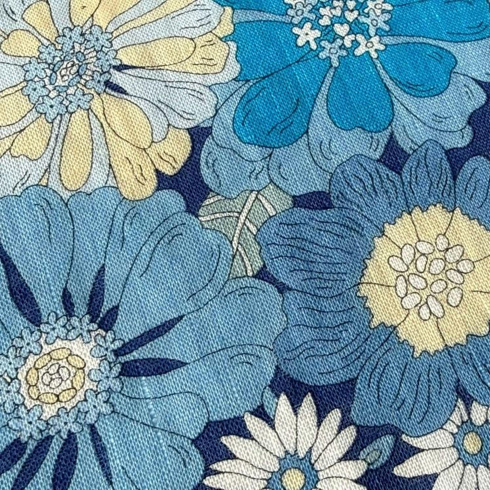 a close-up view of a fabric with a floral pattern in shades of blue and yellow. The fabric appears to be a textile, possibly a piece of clothing or a decorative item. The pattern consists of various flowers and leaves, with some having blue and yellow petals and others having white petals. The overall design is visually appealing and showcases the intricate details of the floral pattern.