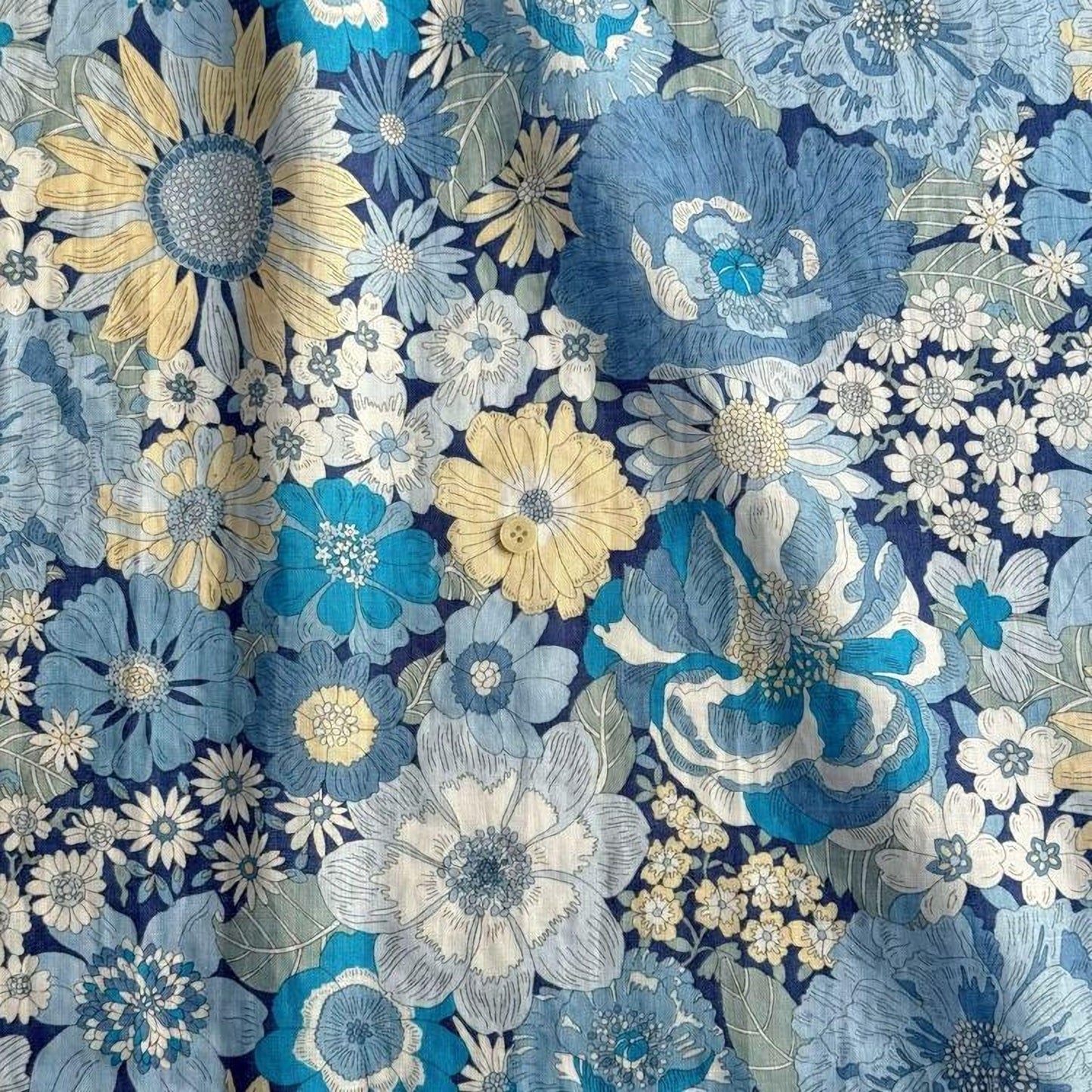 The image is a close-up of a fabric with a vibrant floral pattern in shades of blue, yellow, and white. The pattern features various types of flowers, including sunflowers, daisies, and other blooms, arranged in a visually appealing manner. The fabric appears to be a textile or material with a floral design, likely used for clothing or home decor.