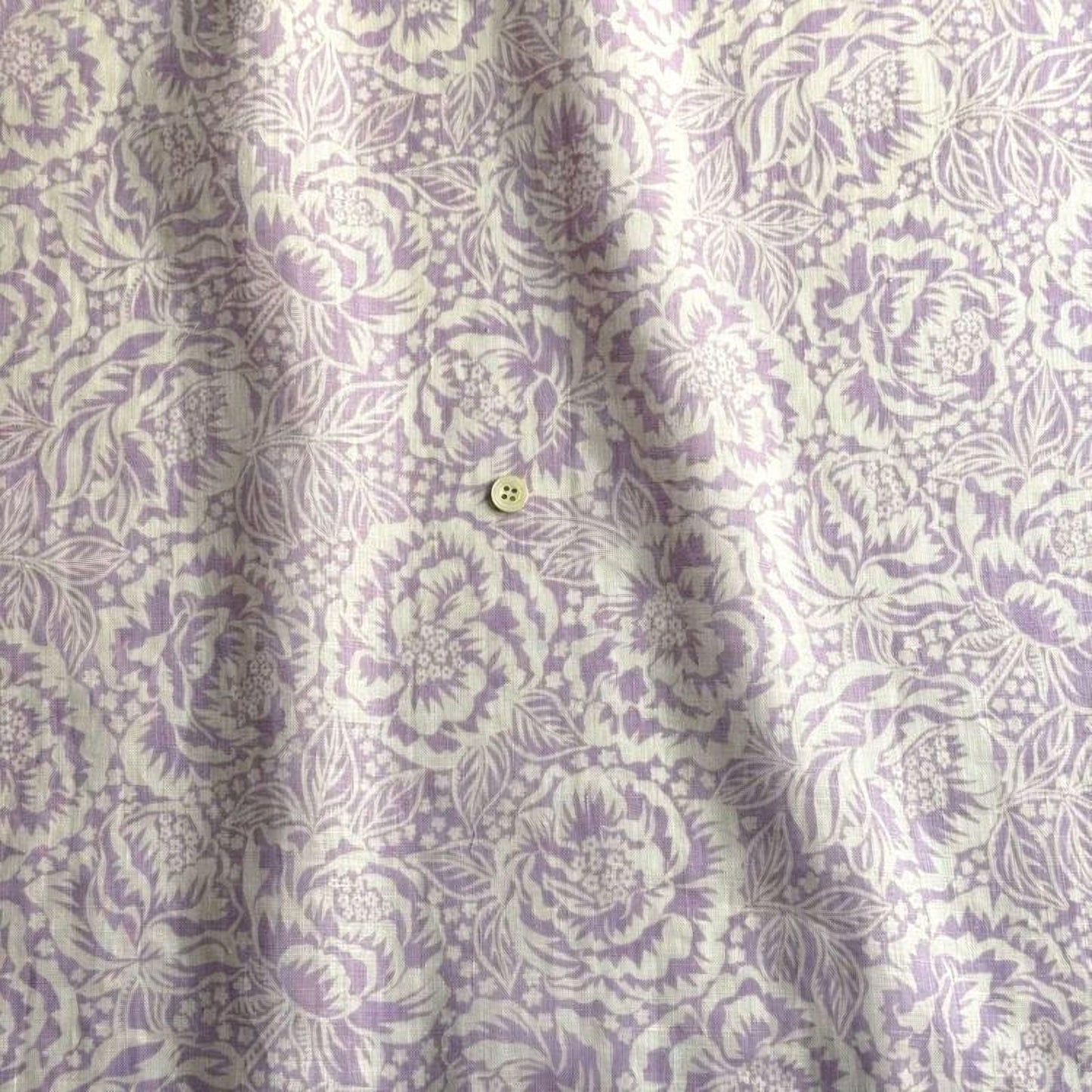 a close-up view of a fabric with a repeating floral pattern in shades of purple and white. The fabric appears to be made of a soft, textured material, and there is a small button visible in the center of the fabric.