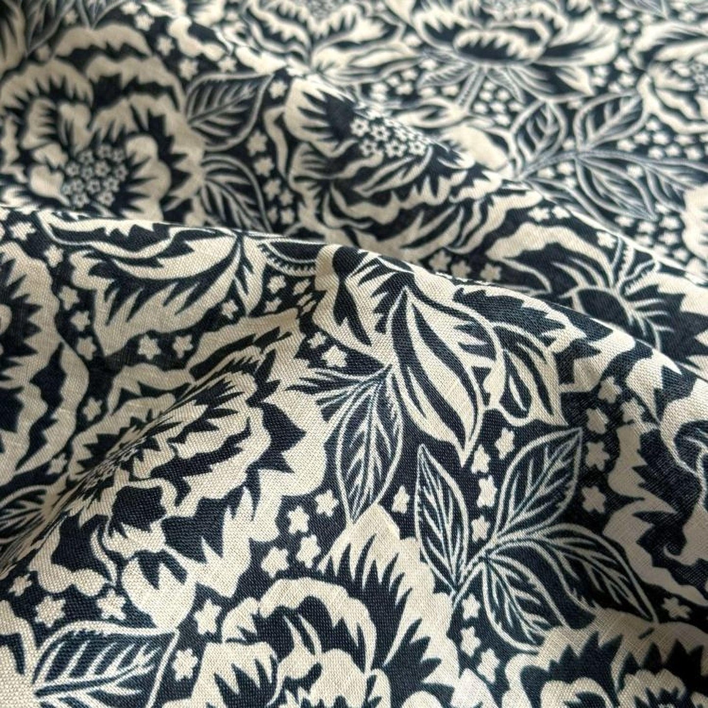 a close-up view of a fabric with a black and white floral pattern. The fabric appears to be made of a soft, textured material, possibly cotton or linen. The pattern consists of various floral designs, including leaves and flowers, arranged in a repeating, intricate manner. The fabric has a slightly wrinkled appearance, suggesting it is made of a natural, flexible material.