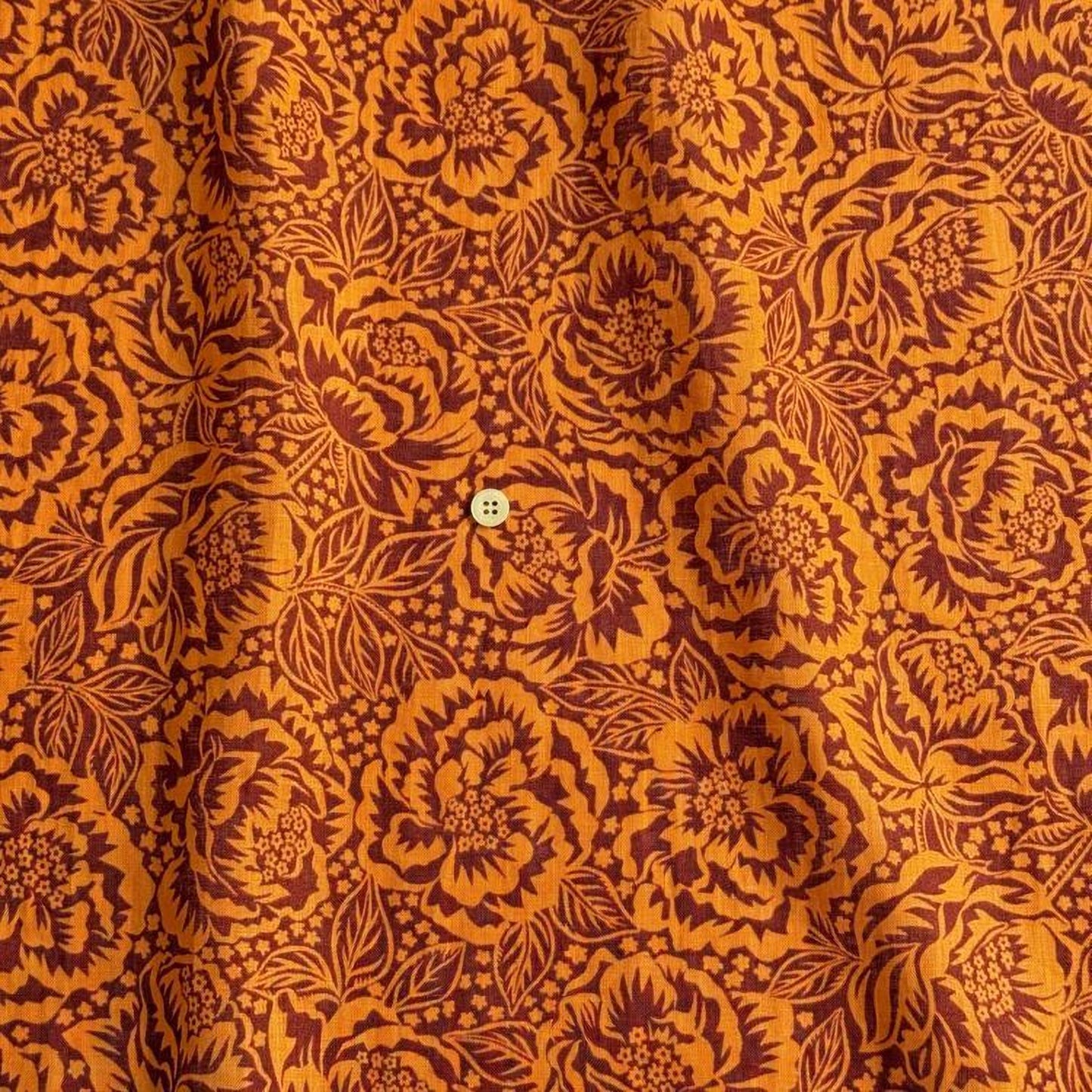 a close-up view of a fabric with a floral pattern in shades of orange and brown. The pattern is intricate and detailed, with numerous flowers and leaves covering the fabric. There is a small button visible in the center of the pattern.