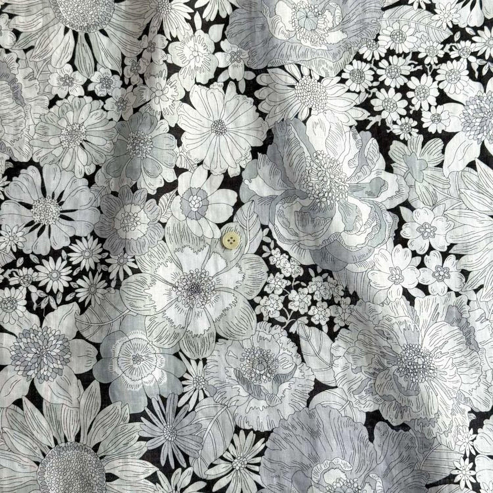 a close-up view of a fabric with a black and white floral pattern. The pattern consists of various flowers and leaves, including sunflowers, daisies, and other types of flowers, all rendered in a monochromatic style. The fabric appears to be made of a material that resembles cotton or linen, and the image does not contain any text or other discernible objects.