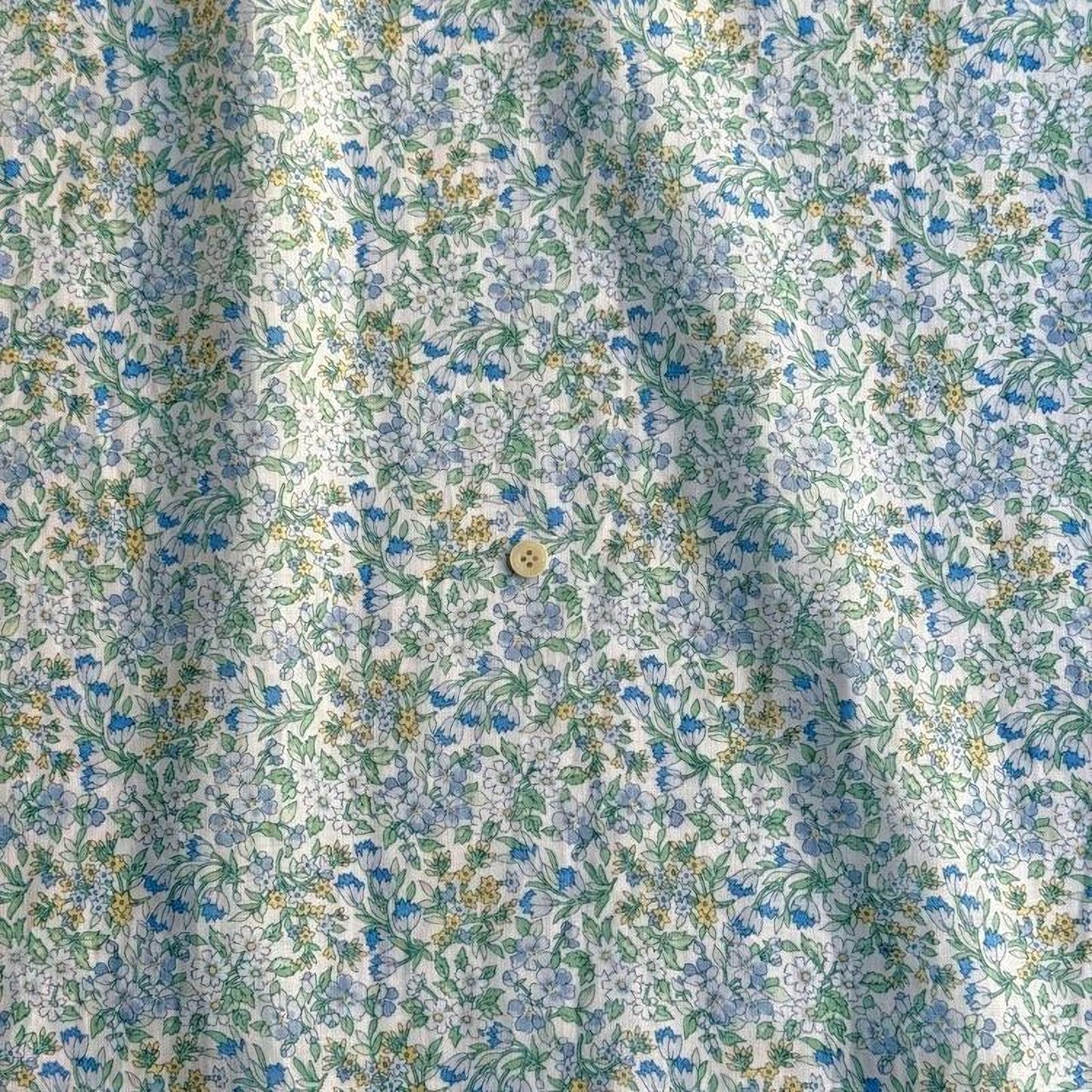 a close-up view of a fabric with a floral pattern in shades of blue, green, and yellow. The fabric appears to be a light-colored material, possibly cotton or linen, with a subtle texture. There is a small button visible in the center of the fabric.