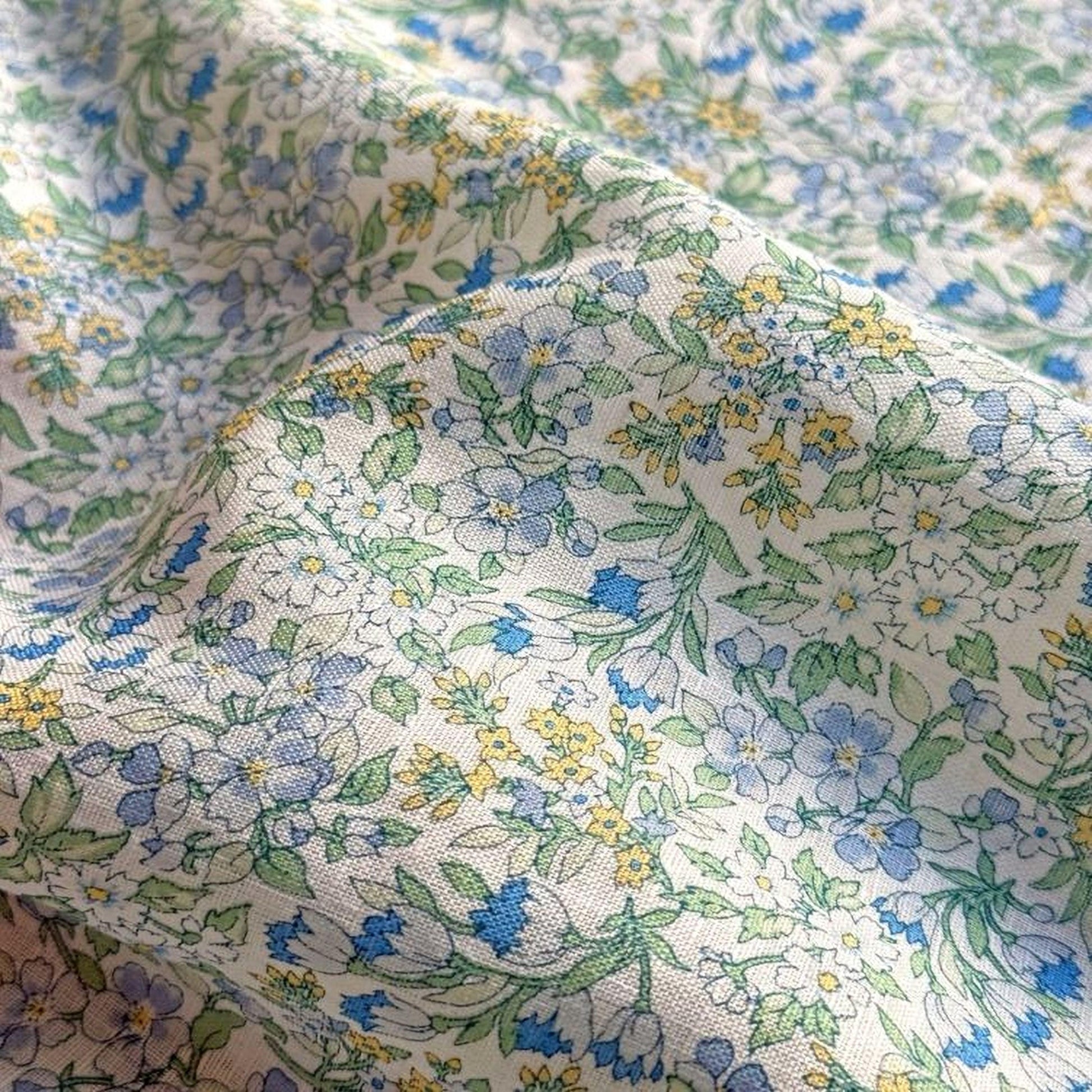 a close-up view of a fabric with a floral pattern in shades of blue, green, and yellow. The pattern is intricate and detailed, with various flowers and leaves interspersed throughout the fabric. The fabric appears to be made of a soft, textured material, and the colors are vibrant and eye-catching.