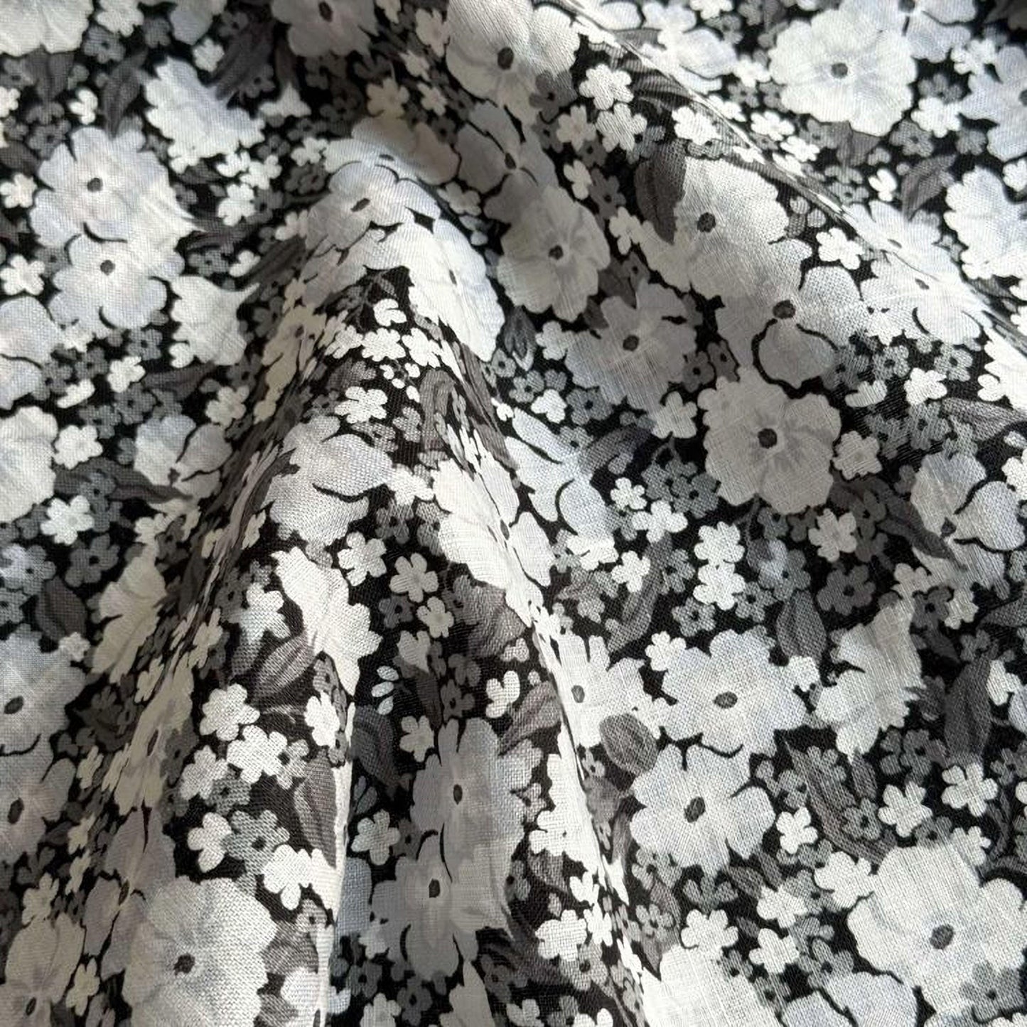 a close-up view of a fabric with a black and white floral pattern. The fabric appears to be made of a soft, textured material, possibly cotton or linen. The pattern consists of numerous small white flowers interspersed with black leaves, creating a visually striking contrast against the dark background. The fabric appears to be folded or crumpled, adding a sense of texture and depth to the image.