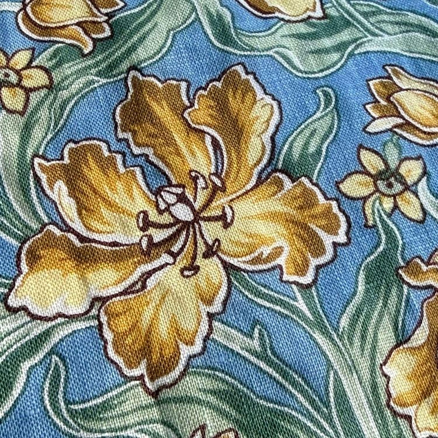 a close-up view of a fabric with a floral pattern. The fabric has a blue background with yellow flowers and green leaves. The flowers are the main focus of the image, with one large yellow flower in the center and smaller yellow flowers surrounding it. The green leaves are scattered throughout the fabric, adding to the overall floral design.