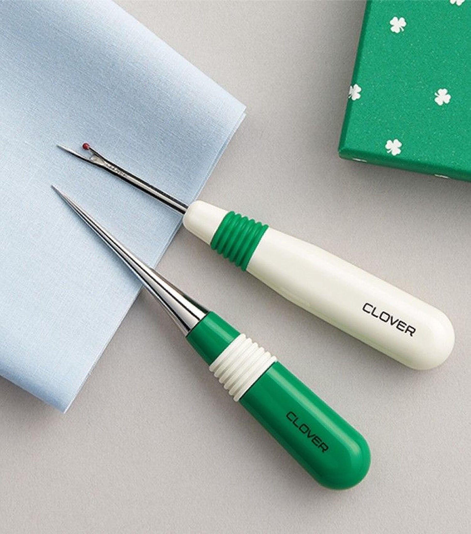 two green and white sewing tools, specifically clover scissors, placed on a light-colored surface alongside a green box with white clover designs.