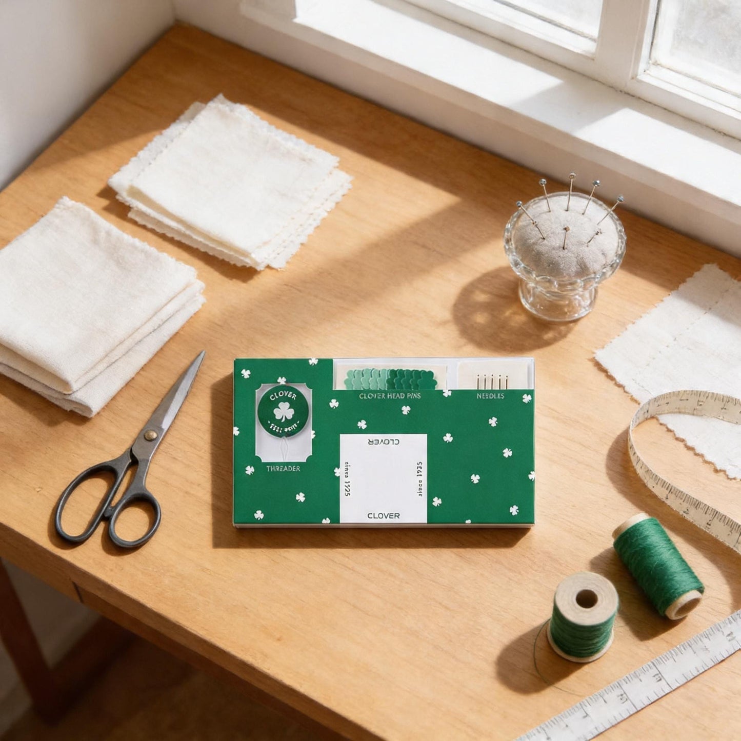 a table with various sewing and crafting supplies, including a green and white package labeled "CLOVER", a pair of scissors, a spool of thread, and a small glass container with pins.