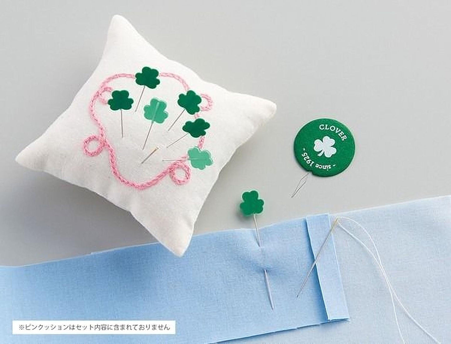 a white pillow with green clover pins and a green pin with the text "CLOVER" on it, along with some sewing materials such as a blue fabric and a needle.