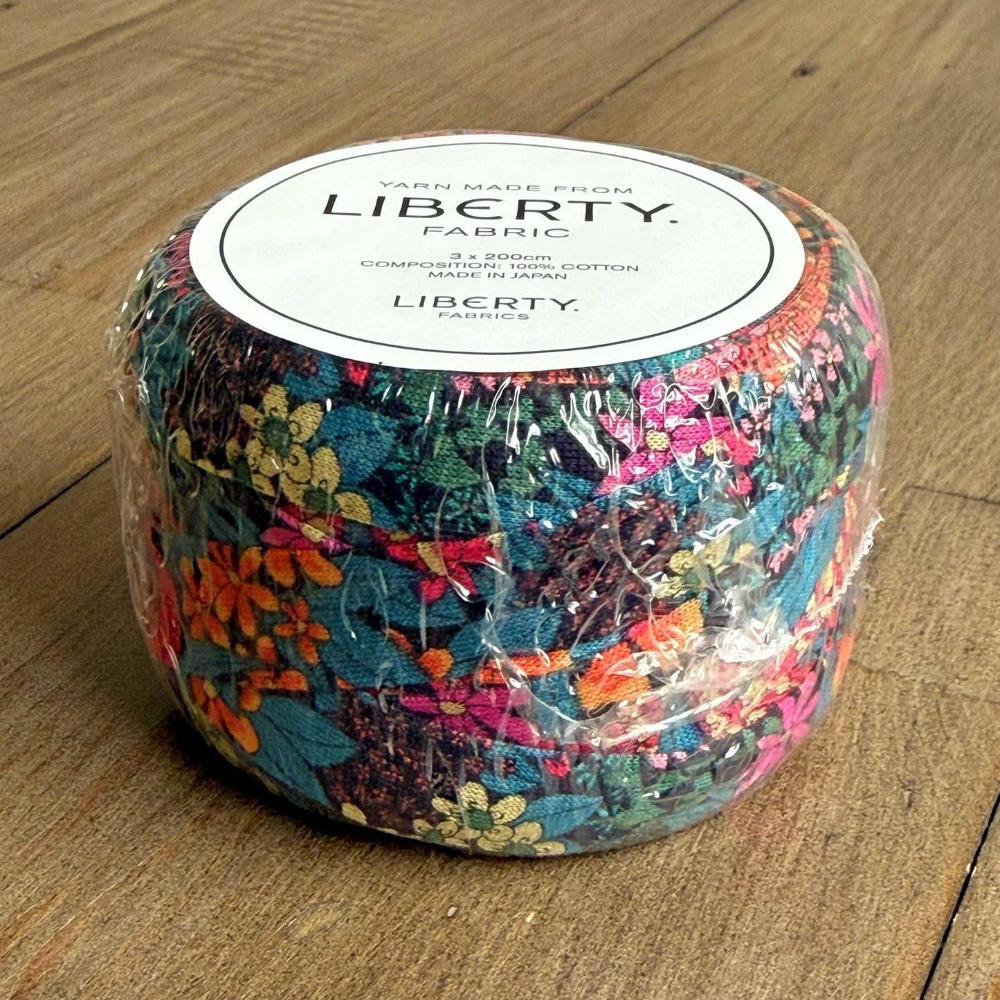 A package of Liberty fabric, featuring a vibrant floral pattern, is wrapped in plastic and placed on a wooden surface.