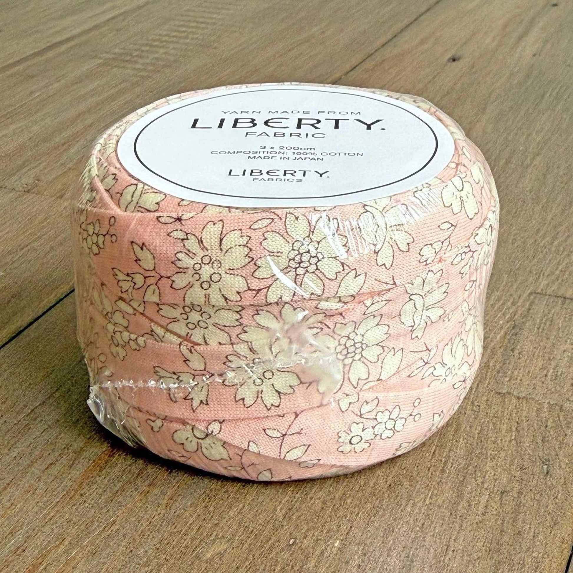 A package of Liberty fabric, which is a pink and white floral patterned fabric, is sitting on a wooden surface.