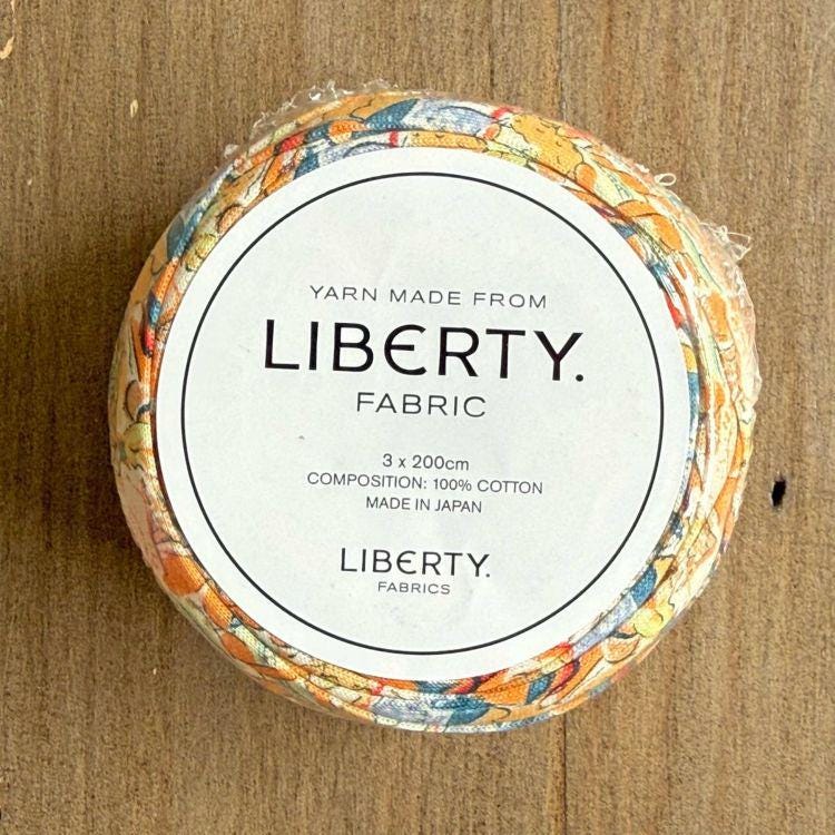 A circular object with a label that reads "Yarn Made from LIBERTY. FABRIC" and "3 x 200cm COMPOSITION: 100% COTTON MADE IN JAPAN".