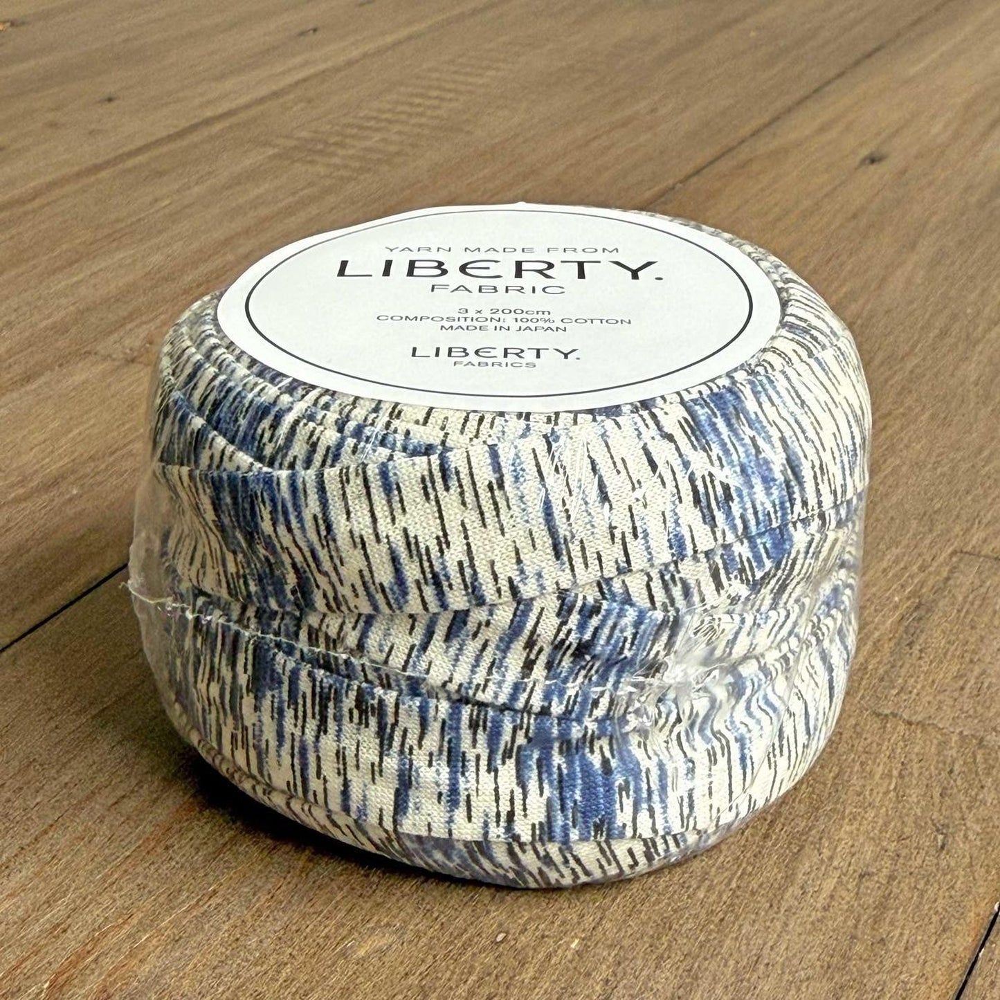 A spool of yarn with a label that reads "Yarn Made from LIBERTY Fabric" is sitting on a wooden surface.