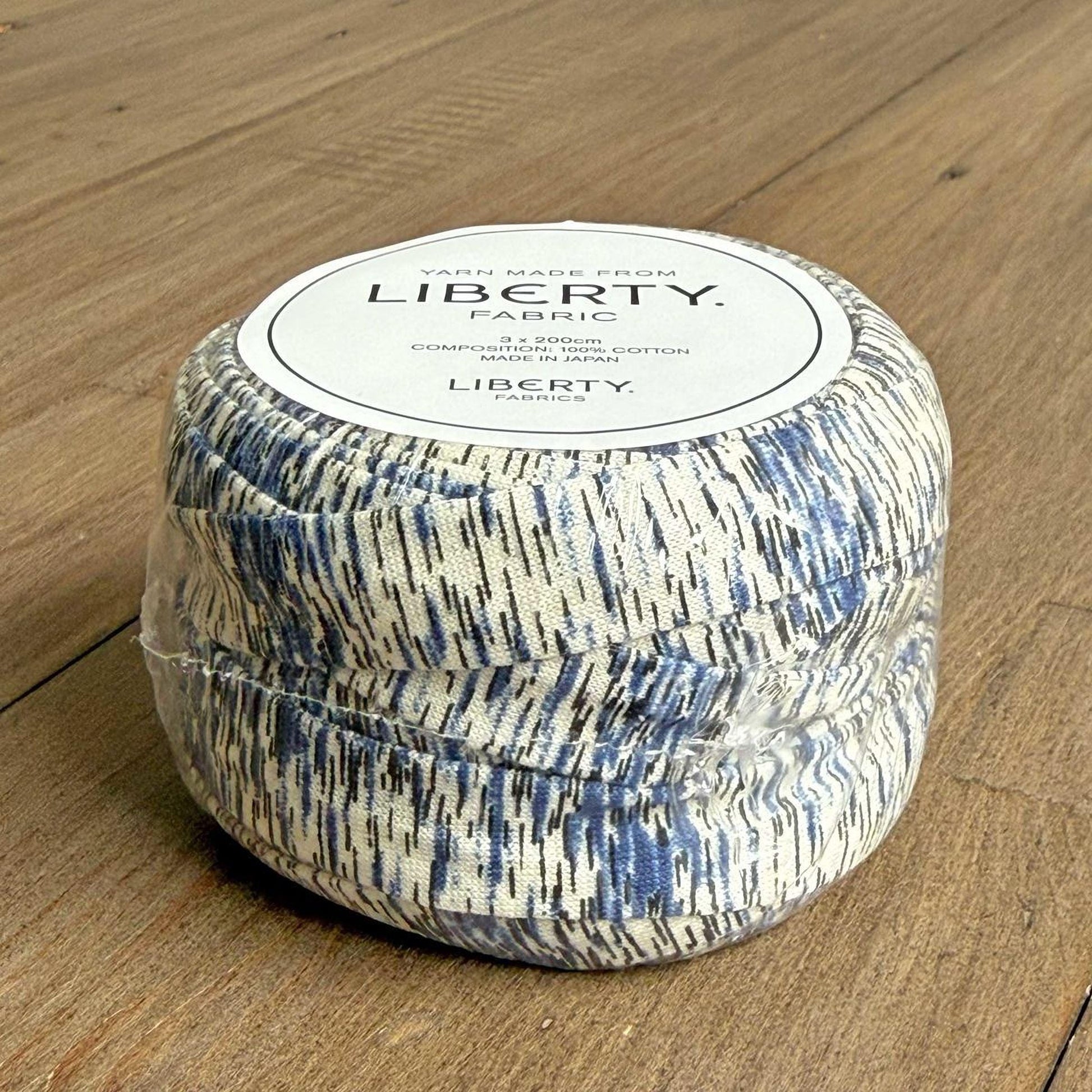 A spool of yarn with a label that reads "Yarn Made from LIBERTY Fabric" is sitting on a wooden surface.