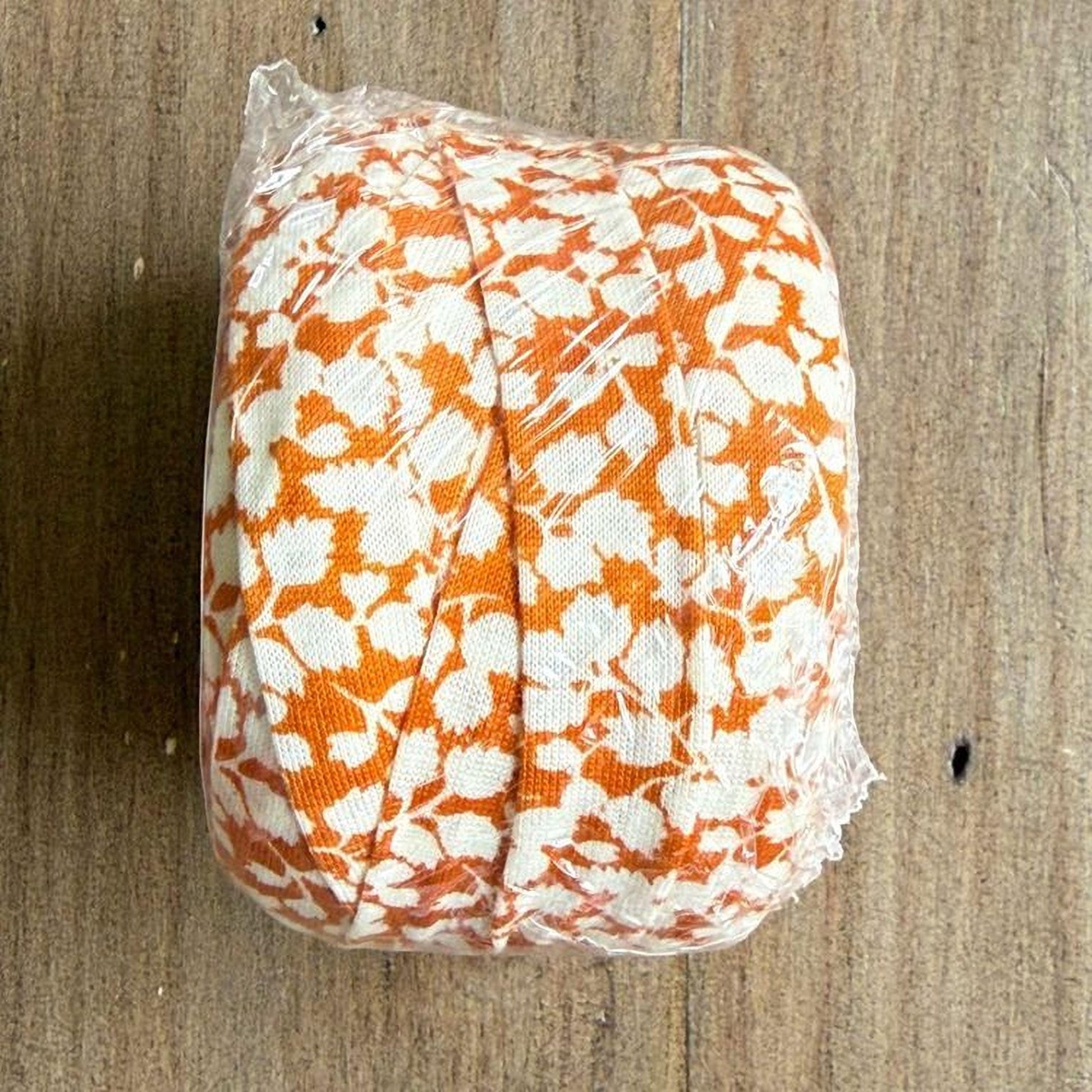 A roll of orange and white fabric with a floral pattern is wrapped in plastic and placed on a wooden surface.