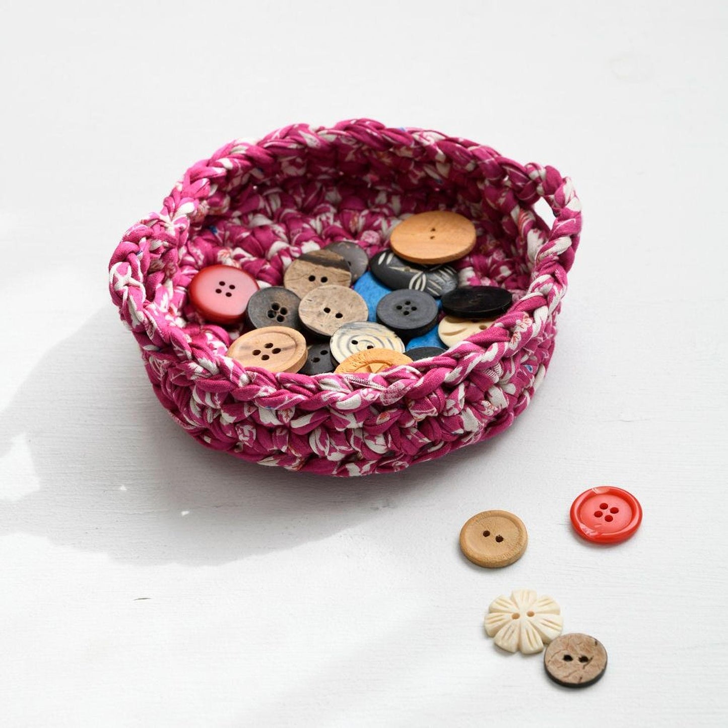 A pink crocheted basket filled with various buttons is placed on a white surface, with some additional buttons scattered around it.