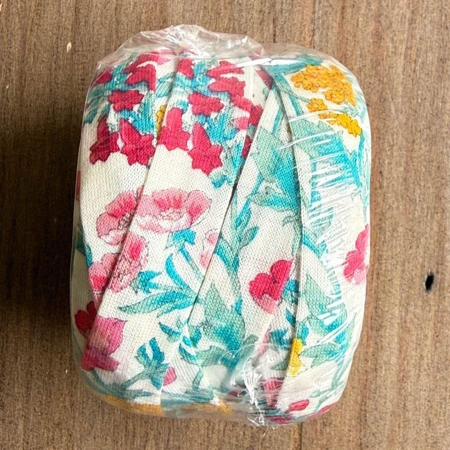 A roll of colorful fabric with a floral pattern is wrapped in plastic and placed on a wooden surface.