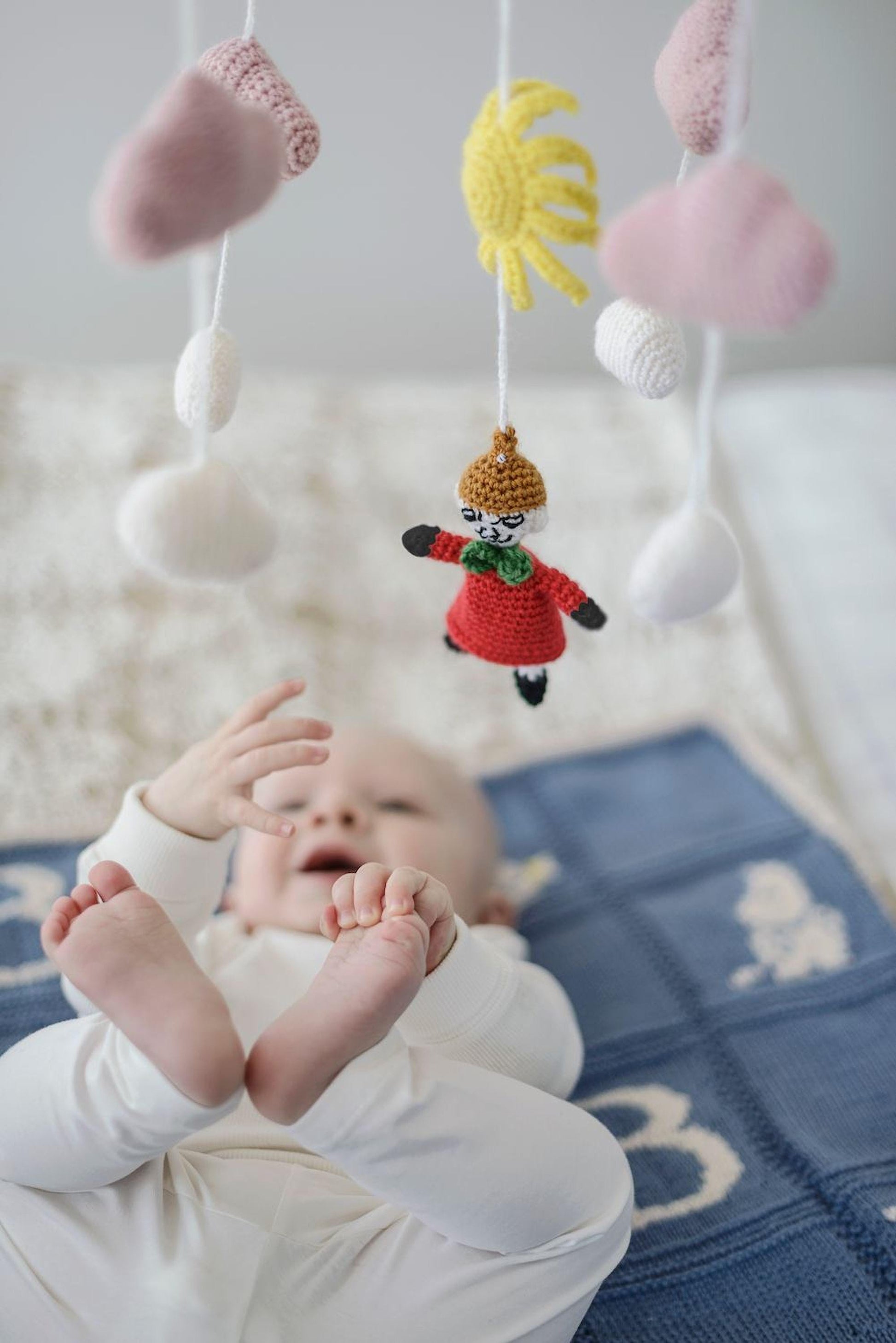 A baby lying on a blue blanket, surrounded by crocheted toys hanging above.