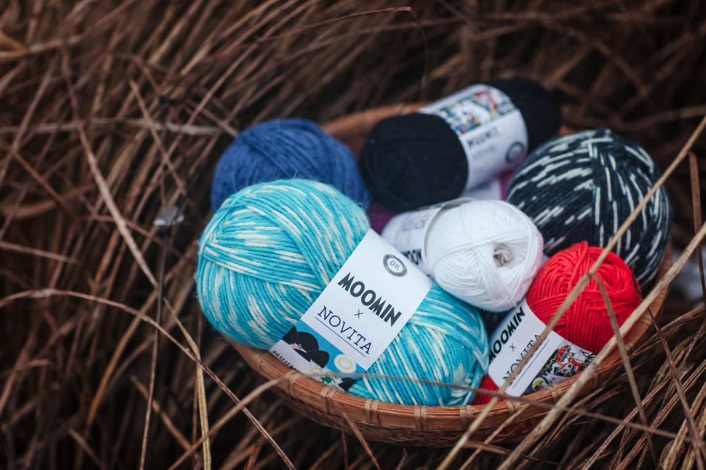 A basket filled with various balls of yarn, including blue, black, white, and red yarn, resting on a bed of straw or grass.