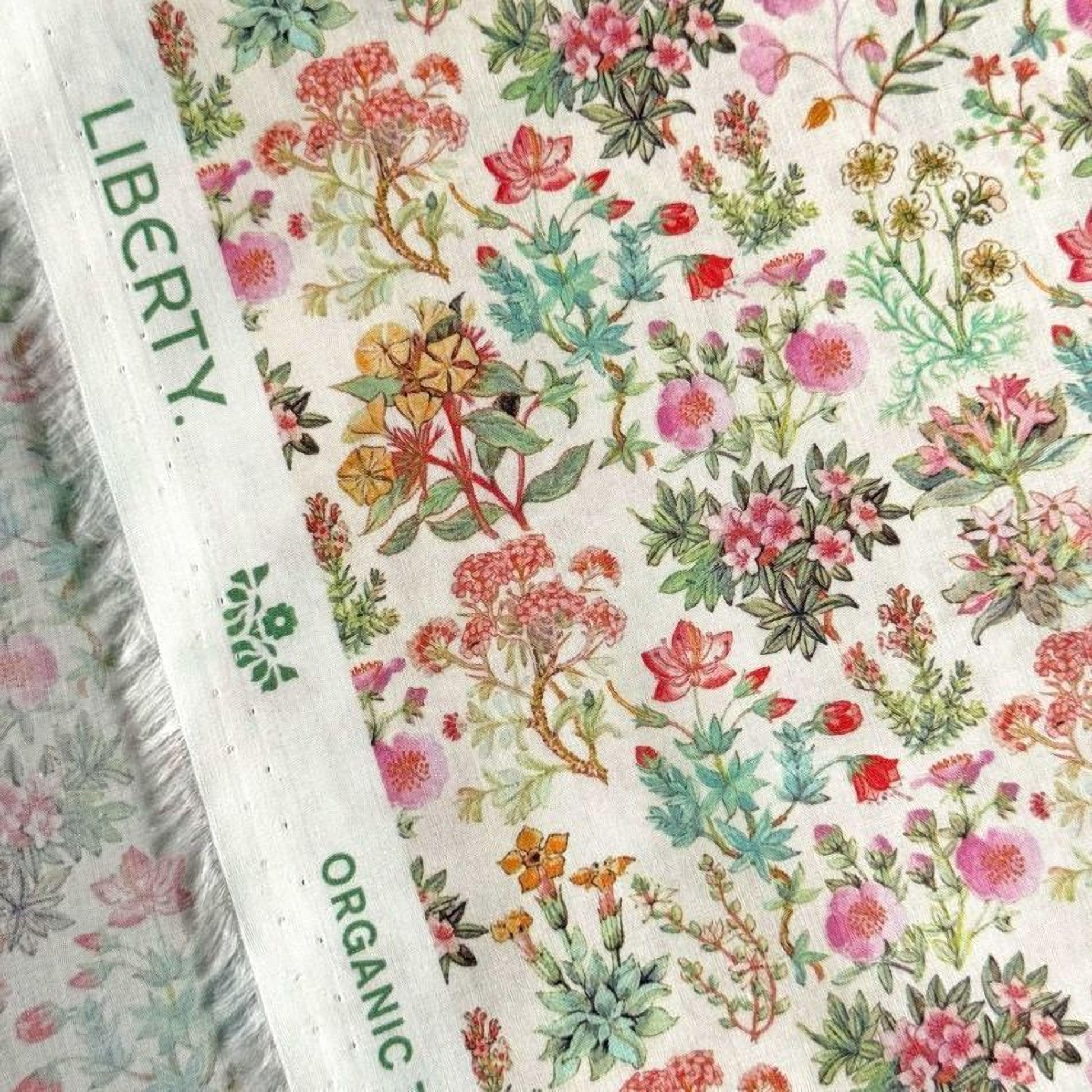 a close-up view of a fabric with a vibrant floral pattern. The fabric appears to be a white material with a variety of colorful flowers and plants printed on it. The text "LIBERTY" and "ORGANIC" can be seen, indicating the brand or type of the fabric.