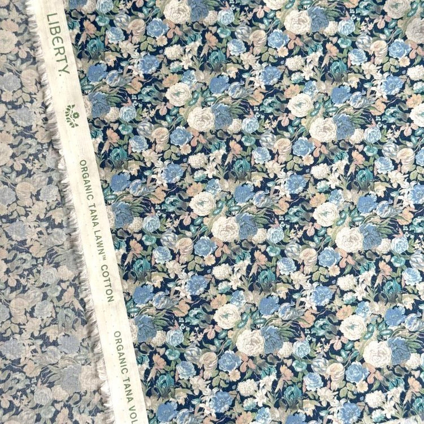 a close-up view of a fabric with a floral pattern, including various shades of blue, green, and white flowers. The fabric appears to be a type of cotton material, as indicated by the text "Liberty" and "Organic TANAL LAWN" visible on the fabric.