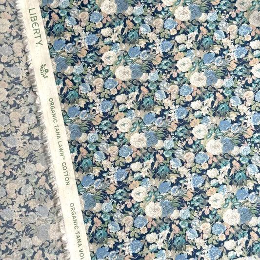 a close-up view of a fabric with a floral pattern, including various shades of blue, green, and white flowers. The fabric appears to be a type of cotton material, as indicated by the text "Liberty" and "Organic TANAL LAWN" visible on the fabric.