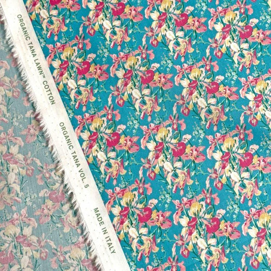 a close-up view of a fabric with a floral pattern in shades of blue and pink. The fabric appears to be made of organic cotton and is displayed on a white fabric with a label that reads "Organic cotton" and "TANA LAWN® VOL. 5".