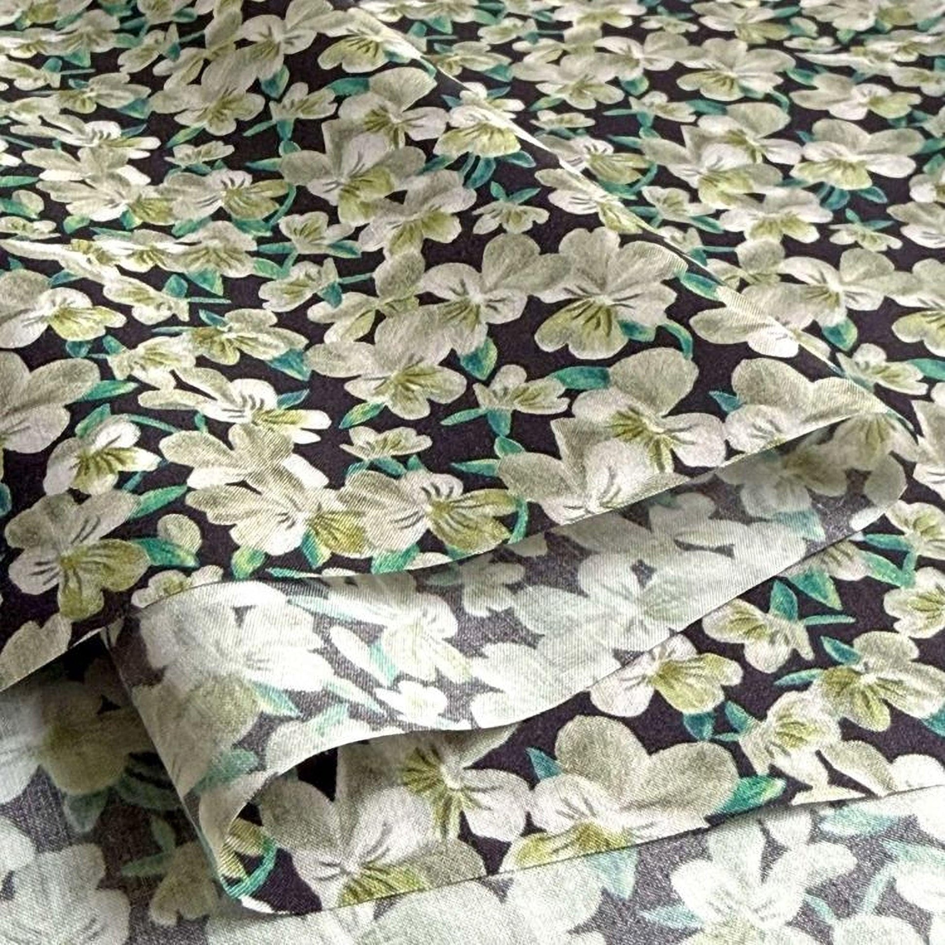 a close-up view of a fabric with a floral pattern in shades of green and white. The fabric appears to be a dark-colored material with a repeating pattern of white flowers and green leaves. The image does not contain any text or other objects, and the focus is solely on the fabric and its floral design.